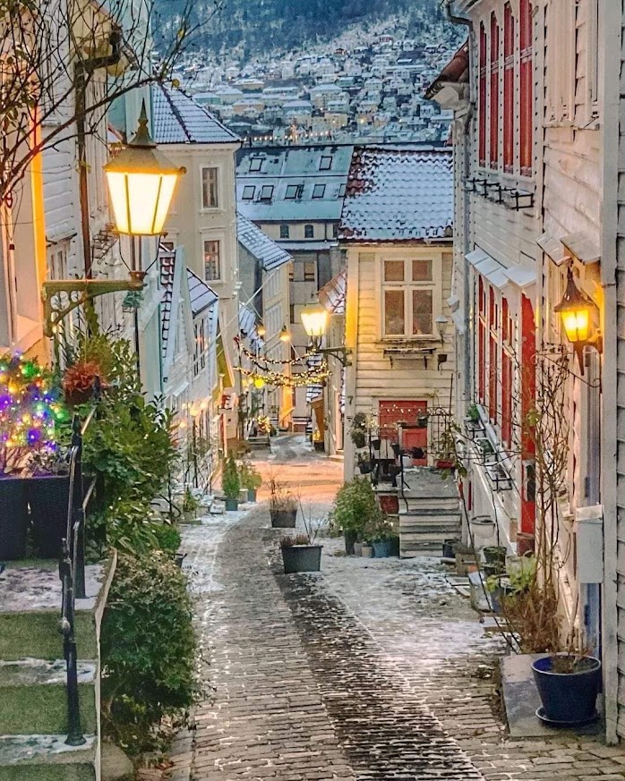 Snow-dusted cobblestone lane lit by warm lanterns, winding between white wooden houses